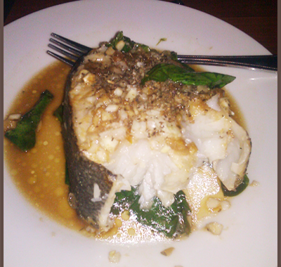 A cooked fish steak on a plate with sauce and greens.